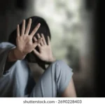 Person sitting against a wall, hands raised to shield their face, appearing distressed.