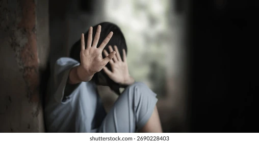 Person sitting against a wall, hands raised to shield their face, appearing distressed.