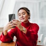 Woman in a red sweater sits at a table, looking at her smartphone and smiling as she takes a selfie.