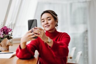 Woman in a red sweater sits at a table, looking at her smartphone and smiling as she takes a selfie.