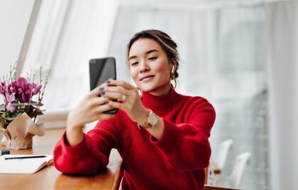 Woman in a red sweater sits at a table, looking at her smartphone and smiling as she takes a selfie.
