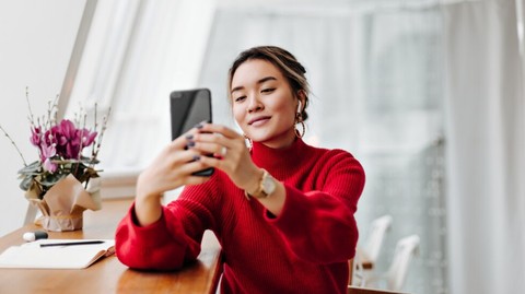 Woman in a red sweater sits at a table, looking at her smartphone and smiling as she takes a selfie.