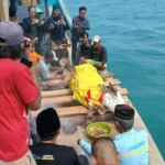A group of men on a wooden boat by turquoise sea photograph a body wrapped in a bright yellow cloth lying on planks, while others record with cameras; a green bowl of offerings sits nearby.