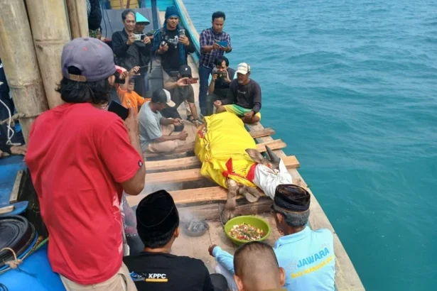 A group of men on a wooden boat by turquoise sea photograph a body wrapped in a bright yellow cloth lying on planks, while others record with cameras; a green bowl of offerings sits nearby.