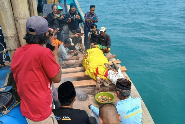 A group of men on a wooden boat by turquoise sea photograph a body wrapped in a bright yellow cloth lying on planks, while others record with cameras; a green bowl of offerings sits nearby.