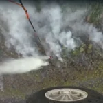 Aerial view of a drone spraying a white mist over a rocky hillside, with a wheel visible in the lower foreground.