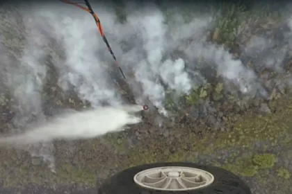 Aerial view of a drone spraying a white mist over a rocky hillside, with a wheel visible in the lower foreground.