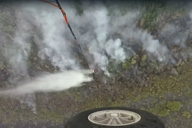Aerial view of a drone spraying a white mist over a rocky hillside, with a wheel visible in the lower foreground.