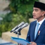 Man in a blue suit and black peci delivering a speech at a podium with three microphones, holding a blue folder.