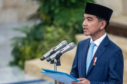 Man in a blue suit and black peci delivering a speech at a podium with three microphones, holding a blue folder.