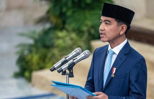 Man in a blue suit and black peci delivering a speech at a podium with three microphones, holding a blue folder.