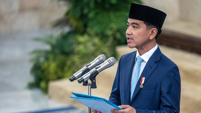 Man in a blue suit and black peci delivering a speech at a podium with three microphones, holding a blue folder.