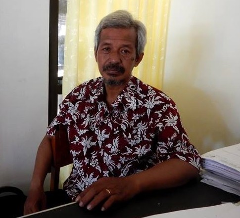 Man with gray hair in a red floral shirt sits at a desk with a stack of papers in a bright room nearby yellow curtains.