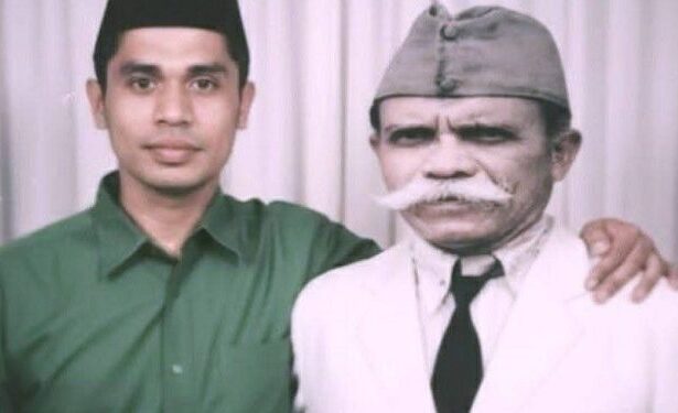 Two men pose for a photo: a younger man in a green shirt with his arm around an older man in a white suit and gray cap.