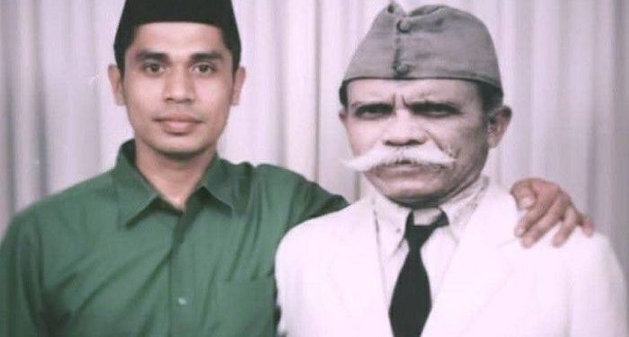 Two men pose for a photo: a younger man in a green shirt with his arm around an older man in a white suit and gray cap.