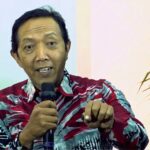 Man wearing a red-patterned shirt speaks into a handheld microphone and gestures toward the viewer.