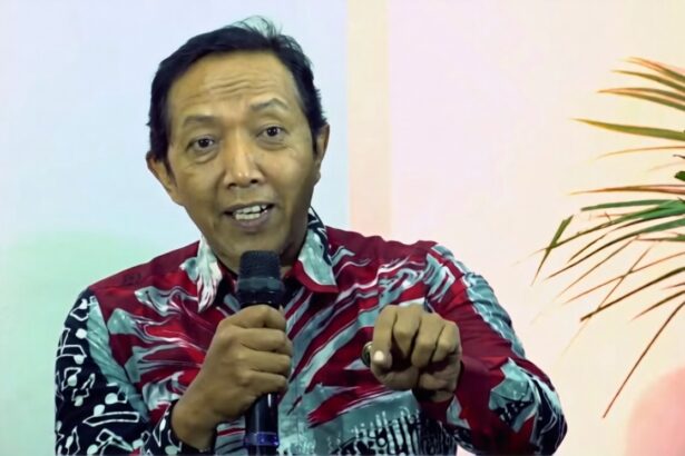 Man wearing a red-patterned shirt speaks into a handheld microphone and gestures toward the viewer.