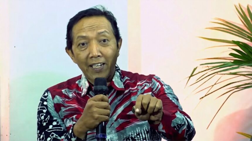 Man wearing a red-patterned shirt speaks into a handheld microphone and gestures toward the viewer.