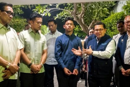 A man in a dark blue shirt gestures as others listen in a garden-like courtyard.