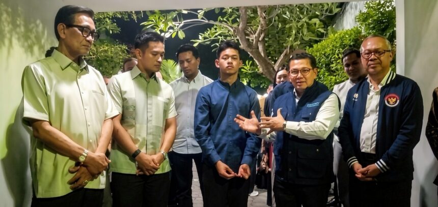 A man in a dark blue shirt gestures as others listen in a garden-like courtyard.