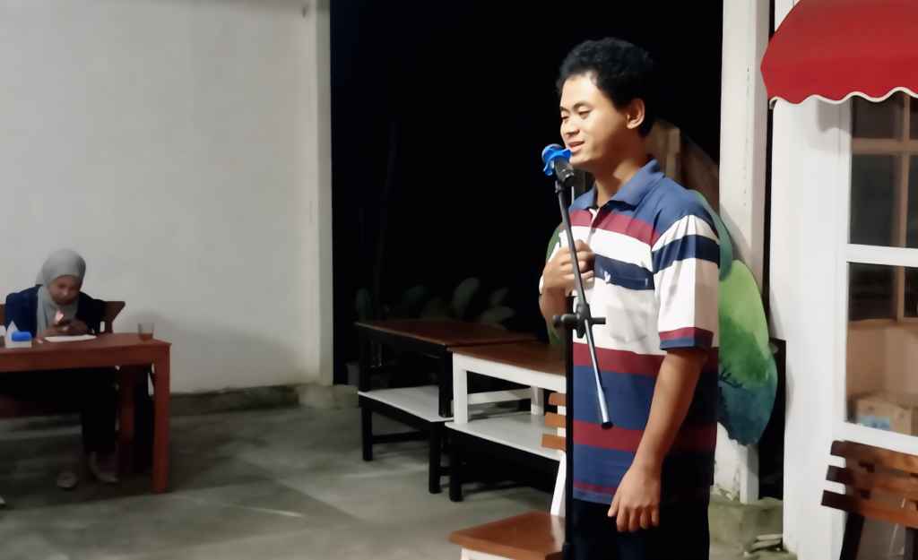 Young man in a striped polo shirt sings into a microphone on a small outdoor stage at night, with a woman in a hijab seated at a desk to the left.