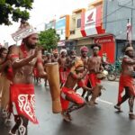Group of dancers in red loincloths and body paint performing with drums on a city street during a parade.