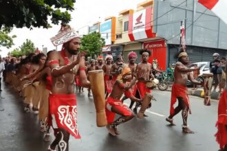 Group of dancers in red loincloths and body paint performing with drums on a city street during a parade.