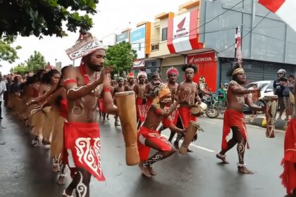 Group of dancers in red loincloths and body paint performing with drums on a city street during a parade.