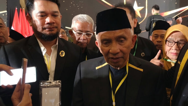 Group of people at a formal event; central older man in a black cap speaks as others photograph him with phones.