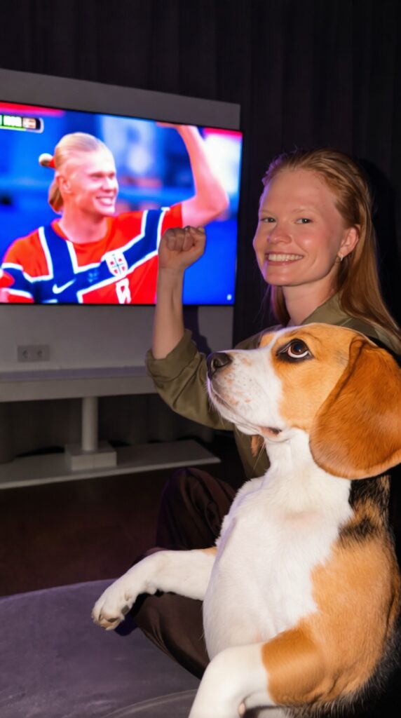 Smiling woman with a beagle watching a sports game on a flat-screen TV in a living room, fist raised in excitement.