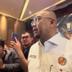 Man in a white shirt with a crest speaks to reporters as several people hold up smartphones to film him at a press event in a hall.