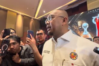 Man in a white shirt with a crest speaks to reporters as several people hold up smartphones to film him at a press event in a hall.