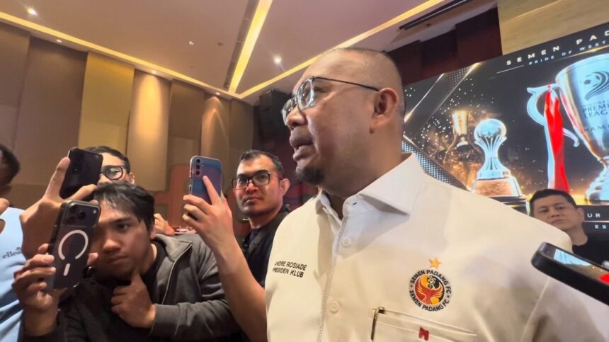 Man in a white shirt with a crest speaks to reporters as several people hold up smartphones to film him at a press event in a hall.