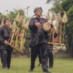 Three elderly performers in black robes walk across a village field, carrying bamboo frames with straw decorations and a drum in hand.