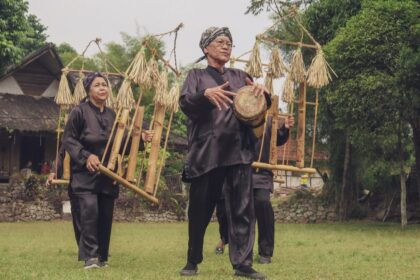 Three elderly performers in black robes walk across a village field, carrying bamboo frames with straw decorations and a drum in hand.