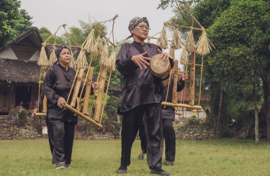 Three elderly performers in black robes walk across a village field, carrying bamboo frames with straw decorations and a drum in hand.