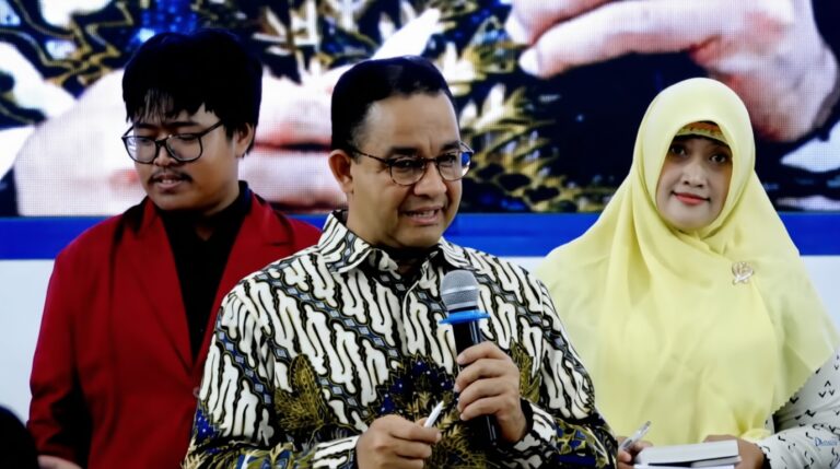 Man in patterned shirt speaks into a handheld microphone at a public event, flanked by a man in a red blazer and a woman in a pale yellow hijab.
