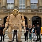 Group of people posing with a large cardboard humanoid sculpture outdoors in front of a historic stone building with arches and banners.