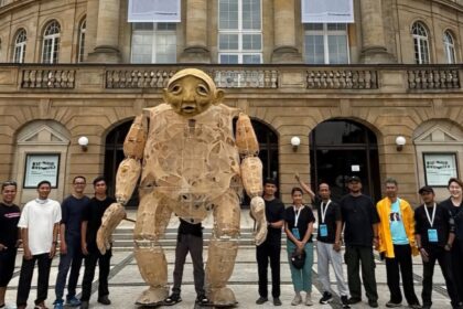 Group of people posing with a large cardboard humanoid sculpture outdoors in front of a historic stone building with arches and banners.
