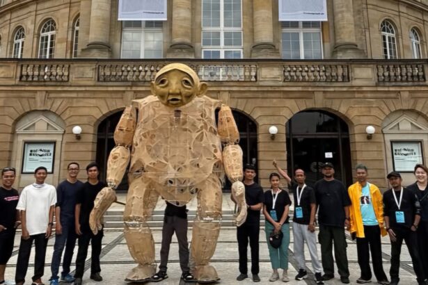 Group of people posing with a large cardboard humanoid sculpture outdoors in front of a historic stone building with arches and banners.
