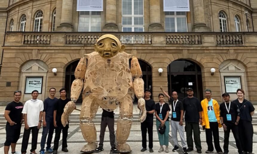 Another Journey with papermoonpuppet Stream of Memory19-20 June 2026Opernhaus Chemnitz Germany 2 - Mabur.co Group of people posing with a large cardboard humanoid sculpture outdoors in front of a historic stone building with arches and banners.