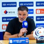 Anthony Hudson, head coach, at a press conference with a blue microphone; a soccer ball sits on the table beside him.