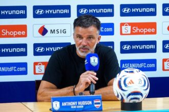 Anthony Hudson, head coach, at a press conference with a blue microphone; a soccer ball sits on the table beside him.