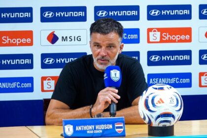 Anthony Hudson, head coach, at a press conference with a blue microphone; a soccer ball sits on the table beside him.