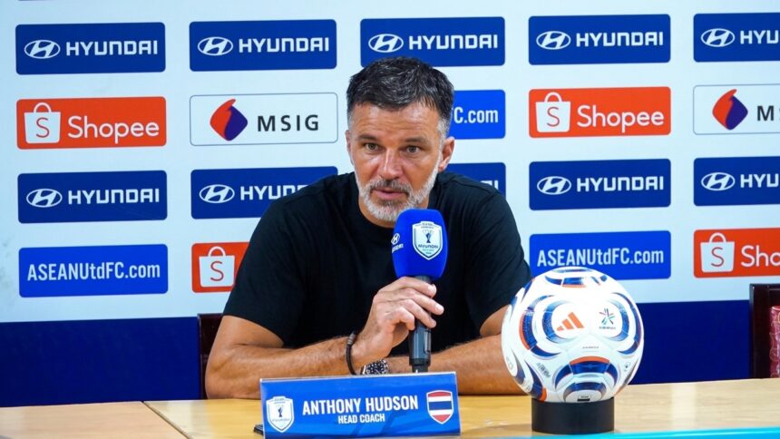 Anthony Hudson, head coach, at a press conference with a blue microphone; a soccer ball sits on the table beside him.