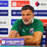 Anthony Hudson, head coach, sits at a press conference table with a microphone, wearing a green shirt; sponsor-backed backdrop in view.