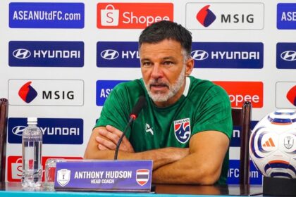 Anthony Hudson, head coach, sits at a press conference table with a microphone, wearing a green shirt; sponsor-backed backdrop in view.