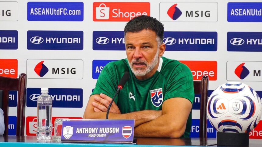 Anthony Hudson, head coach, sits at a press conference table with a microphone, wearing a green shirt; sponsor-backed backdrop in view.