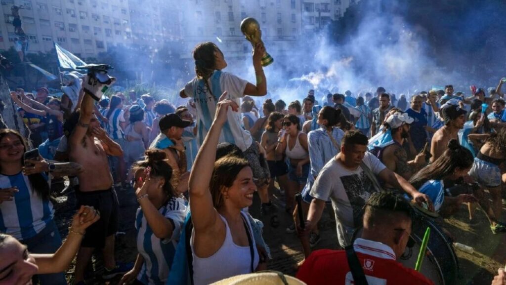 Crowd of fans in blue-and-white jerseys celebrate a victory as a person on another's shoulders raises a trophy amid smoke. It’s a festive sports celebration.