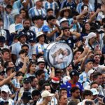 Large cheering crowd in blue-and-white jerseys; a person holds a round drum featuring a kissing couple at center.